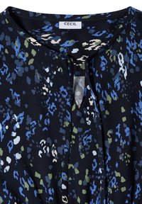 Blouse bleu foncé avec motif abstrait en vert, blanc et bleu, avec un col rond et une petite fente sur le devant ainsi qu'une étiquette de marque Cecil.