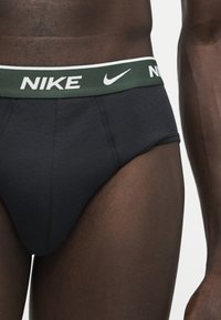 Homme portant un slip Nike noir avec une ceinture verte présentant le logo blanc Nike et le swoosh, visible de la taille jusqu'au haut des cuisses.