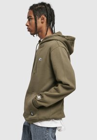 Oliwkowo-zielona bluza z kapturem z kieszenią kangurka, sznurkiem do regulacji kaptura oraz wyszywanym logo na piersi i rękawie. Charakteryzuje się miękką teksturą.