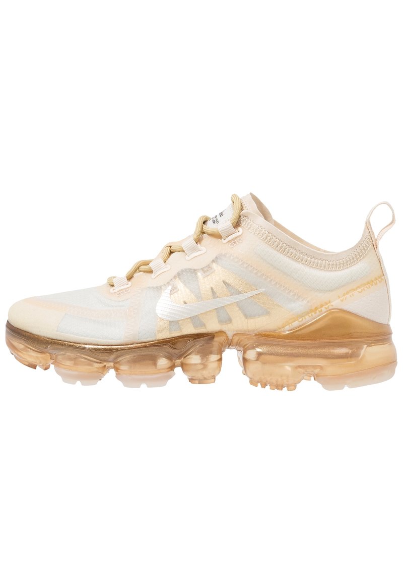 Chaussure de sport beige avec une tige semi-transparente, dotée d'un design texturé, d'accents dorés et d'une semelle claire et rembourrée.
