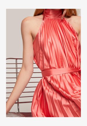 Femme portant une robe corail plissée en satin à col haut, nouée à la taille, avec de longues boucles d'oreilles à pompons, assise sur une chaise en métal.