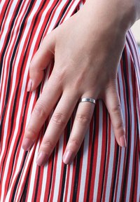 Zilveren ring op een hand met een gladde textuur, rustend op een rood-wit gestreepte stof met dunne zwarte lijnen. Nagels zijn lichtroze.
