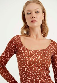 Långärmad topp i rost med vitt bladmönster. Har fyrkantig halsringning och ribbad textur, vilket ger en figurnära silhuett.