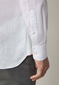 Witte linnen shirt met een gladde textuur, knoopmanchetten en een ronde zoom. Zichtbare roze accent aan de zijnaad, gecombineerd met grijze broek.