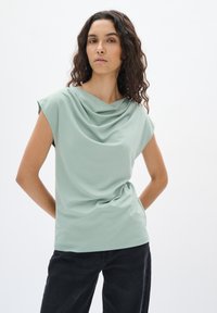 Top verde chiaro a maniche corte con scollo drappeggiato e texture liscia, abbinato a jeans neri. Design semplice e moderno.