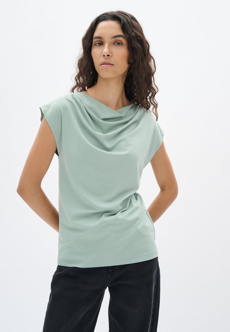 Top verde chiaro a maniche corte con scollo drappeggiato e texture liscia, abbinato a jeans neri. Design semplice e moderno.
