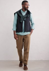 Gilet noir sur une chemise à carreaux verts, pantalon beige et chaussures marron. Présente un design décontracté avec une texture douce et des boutons visibles.