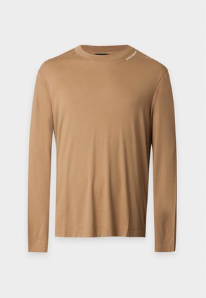Un homme se tient debout portant une chemise marron à manches longues, un jean bleu clair ample et des baskets blanches sur un fond blanc uni.