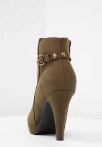 Bottine en suede vert olive avec un talon haut, présentant une sangle décorative avec des clous en forme de pyramide et une semelle noire lisse.