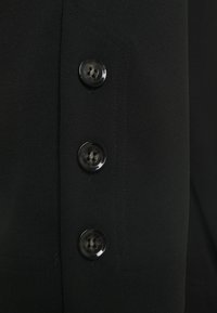 Svart tyg med en slät textur som har tre mörka knappar, var och en med ett centralt hål och en subtil lyster. Skräddarsydd design med sömdetaljer.