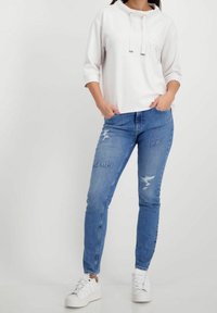 Felpa bianca corta con cappuccio regolabile. Jeans in denim strappati di colore azzurro con la scritta "DENIM" e "ART". Sneakers bianche completano l'outfit.
