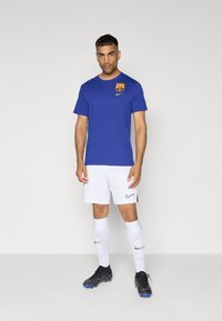 Plava pamučna majica s logotipom FC Barcelone i Nike swoosh, u kombinaciji s bijelim šortsevima, bijelim čarapama do koljena i crnim kopačkama.