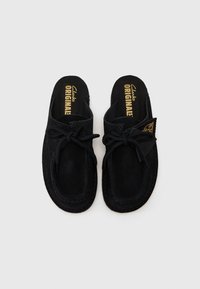 Des chaussures noires en daim à enfiler présentent une forme arrondie, un dessus texturé et un nœud décoratif. La marque est visible sur les semelles intérieures. Des détails cousus sont présents.