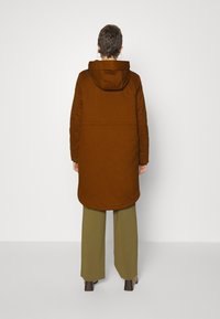 Brun huva parka med en lång, avslappnad passform. Har en mjuk textur och en rundad nederkant. Kombineras med olivgröna vida byxor.