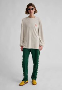 T-shirt de algodão bege de mangas longas com um pequeno logotipo vermelho, combinado com calças de treino verdes com uma risca branca. Calçado: ténis amarelos.