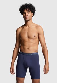 Izmos férfi göndör hajjal, sötétkék Puma boxer sortot viselve, sima világosszürke háttér előtt áll.