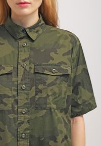 Grüne Camouflage-Hemd mit kurzen Ärmeln, Knöpfen auf der Vorderseite, zwei Brusttaschen und einem Logopatch auf der Tasche. Hergestellt aus leichtem Stoff.