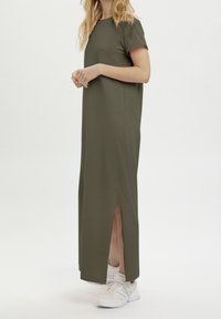 Olivegrünes Maxikleid mit kurzen Ärmeln, seitlichem Schlitz, glatter Textur und lässiger Passform. Trägt weiße Sportschuhe.