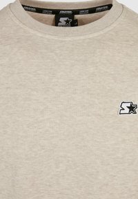 Sudadera beige con cuello redondo, tela texturizada y un pequeño logo negro con una estrella en la zona izquierda del pecho.