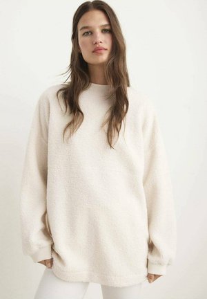 Cream-colored, oversized fleece sweatshirt with a rounded neckline, ribbed cuffs, and a textured surface. Smooth fabric detail is visible.