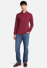 Maglia polo rossa a maniche lunghe con colletto, abbinata a jeans di denim blu e scarpe marroni con lacci, in piedi davanti a uno sfondo semplice.