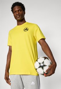 Geltona trumpomis rankovėmis marškinėliai su juodu logotipu, derinami su pilkais sportiniais džinsais. Asmuo laiko baltą futbolą su juodno žvaigždės dizainu.