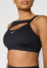 Fekete sportmelltartó kivágott mintával. Rugalmas anyagból készült, kis fehér Nike logóval és széles pántokkal. Simább textúrájú.