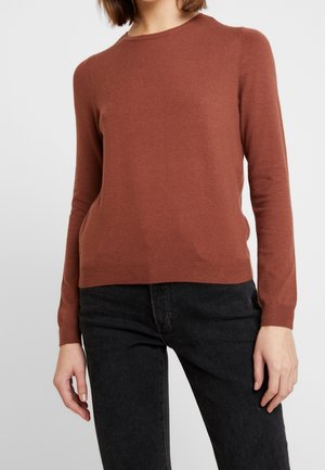 Femme portant un pull à manches longues couleur rouille et un jean noir, debout devant un fond uni gris clair.