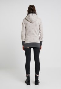 Kvinna som står vänd bort, iförd en beige hoodie med prickar, mörkgrå ärmslut och nederkant, svarta skinny jeans och svarta ankelstövlar.