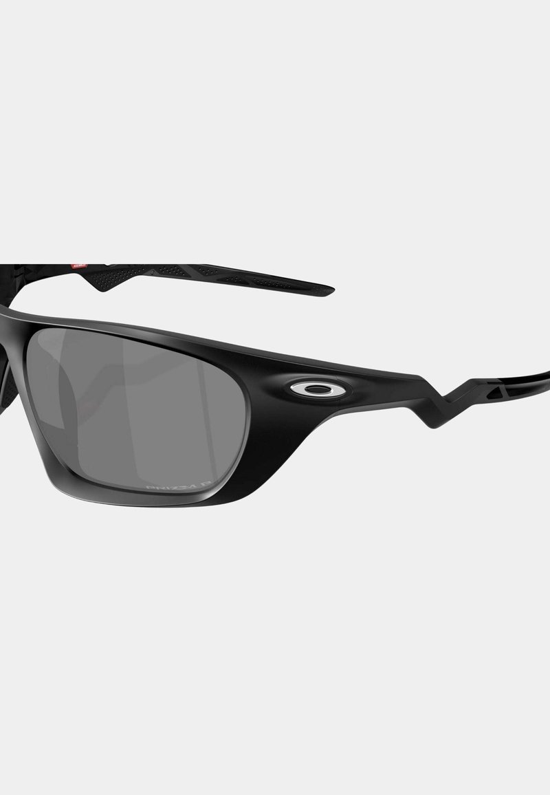 Lunettes de soleil noires avec des verres gris, présentant un design enveloppant et des bords angulaires. Comprend un logo ovale au niveau de la branche et des branches texturées.