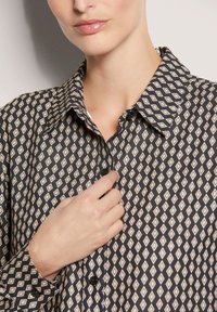 Langärmliges Hemd in Schwarz mit einem Rautenmuster in Beige und Weiß. Es hat einen spitzen Kragen und Knöpfe entlang der Vorderleiste.