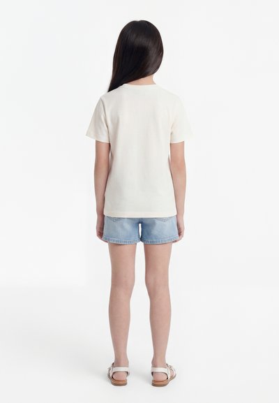 Child with long dark hair wearing a plain white short-sleeve shirt, light blue denim shorts, and white sandals, standing facing away.