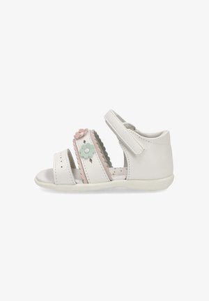 Sandalia de cuero blanca con múltiples correas, bordes en forma de onda y flores decorativas en rosa y verde. Suela de color claro con fondo texturizado.