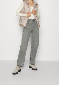 Chaleco acolchado ligero con capucha en beige suave, combinado con jeans grises de talle alto y corte relajado con dobladillos sin terminar, y zapatos beige con cordones y suelas negras.