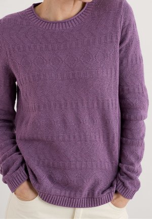 Personne portant un pull en maille texturée violet avec des motifs géométriques, mains dans les poches d'un pantalon couleur crème, sur un fond neutre.