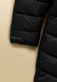 Juodas pūkinis striukės rankovės skyrius su siuvinėtais skyriais ir "JOULES" logotipu, atspausdintu netoli rankogalių, smėlio spalvos fone.