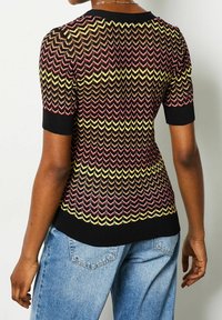 Pletený top s cik-cak vzorem v červené, žluté a černé barvě, s krátkými rukávy a přiléhavým střihem. Texturovaná tkanina s žíhanými lemy.
