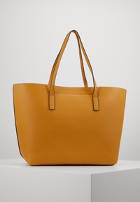 Grande borsa tote in pelle gialla senape texturizzata con due lunghe maniglie, posizionata su un piedistallo bianco contro uno sfondo grigio chiaro semplice.