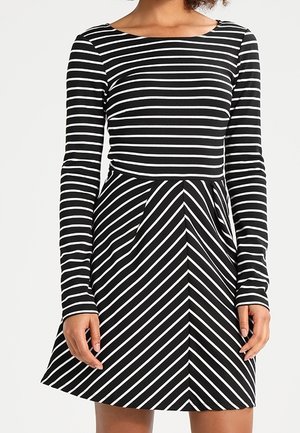 Femme portant une robe noire à manches longues, avec des rayures horizontales sur le haut et des rayures diagonales sur la jupe.