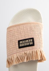 Slide sandal featuring a woven pink upper with fringe, a black logo patch, and a white textured sole.