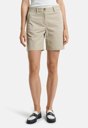 Shorts en coton beige avec un devant plat, deux poches et une fermeture à bouton. Texture lisse, longueur au genou, accompagnés de mocassins foncés.