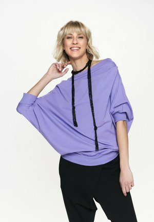 Fioletowy, oversize'owy sweatshirt z rękawami typu dolman, z czarnym frędzlem przy dekolcie. Noszony z czarnymi spodniami. Gładki materiał.