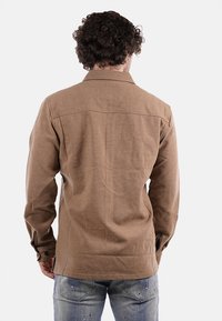 Camicia marrone con texture, colletto, maniche lunghe e polsini con bottoni, abbinata a jeans azzurri chiari. La camicia presenta uno yoke posteriore standard.