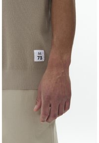 Béžový pletený svetr s žebrovaným lemem; obsahuje bílou etiketu s "M 73." Ruka je umístěna blízko spodní části oděvu.