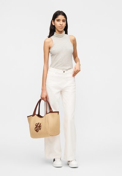 Νεαρή γυναίκα που φοράει ριγέ ζιβάγκο χωρίς μανίκια και λευκό παντελόνι, κρατώντας μια μπεζ πλεκτή τσάντα χειρός με καφέ δερμάτινα χερούλια.