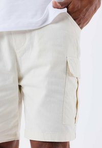 Bermudas cargo de color beige claro, hechas de una tela texturizada, con bolsillos laterales y detalles de costura. Los acabados del dobladillo son limpios, lo que proporciona una forma estructurada.