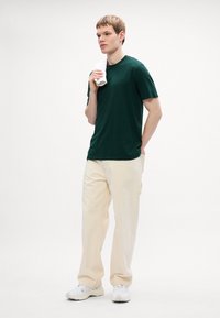Tamnozelena pamučna majica, opuštenog kroja, uparena s ivory oversized hlačama. Model drži bijeli ručnik, nosi bijele tenisice. Minimalistička pozadina.