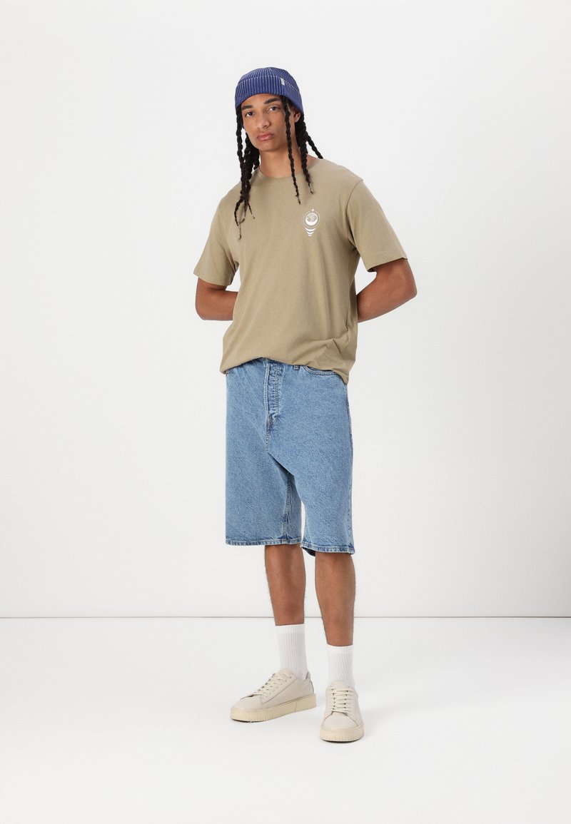 T-shirt kaki à manches courtes avec un logo, shorts en denim bleu, baskets claires et chaussettes blanches. Le modèle a des cheveux tressés et une casquette rayée.