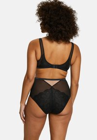 Vrouw met krullend haar gezien van achteren, gekleed in zwarte doorschijnende kanten high-waisted onderbroek en een bijpassende bh tegen een effen achtergrond.