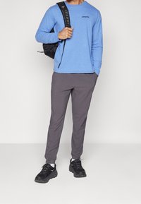 Långärmad blå skjorta med svart logotyp, grå byxor och svarta sneakers. Modellen håller en svart ryggsäck med axelremmar som är synliga.